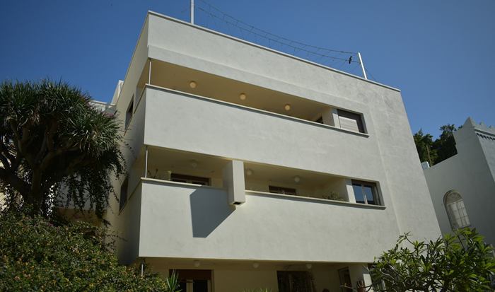 בניין מגורים לבן בסגנון באוהאוס עם מרפסות ארוכות, בין עצים ושמיים כחולים